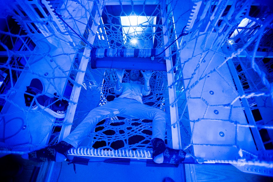 Attendee at an art event. The image is tinted blue. He is dressed in light pants and shirt. He has removed his shoes. He is lying in a braided rope structure that is suspended in the air and playing a keyboard.