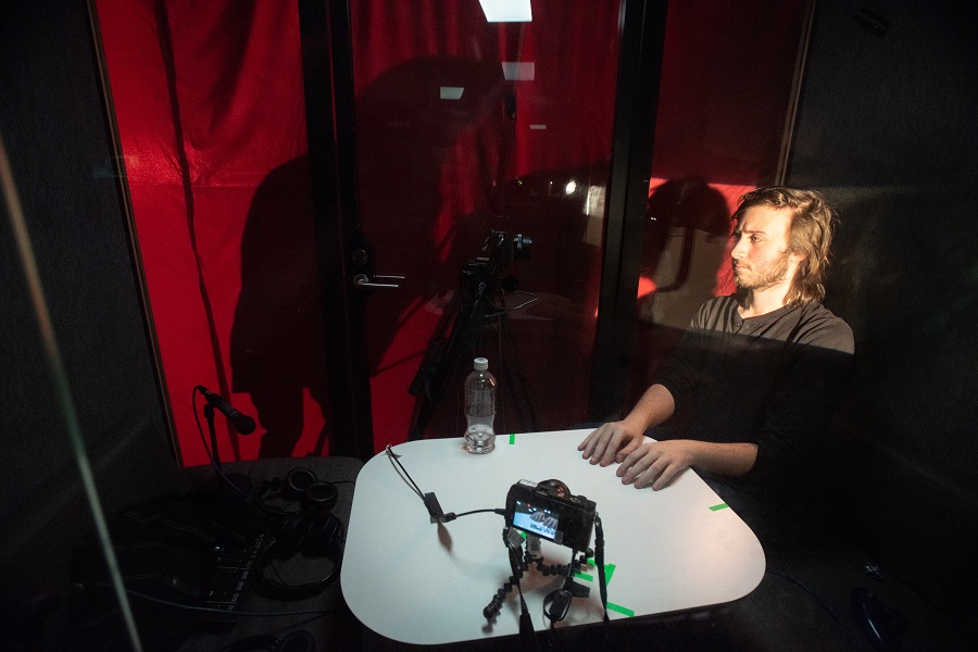 A man in a black shirt sits with his hands on a white surface. The surface has a camera on a tripod pointed at him. His face is framed with light. There is a red curtain behind him and the shadows of two people on the other side are visible.