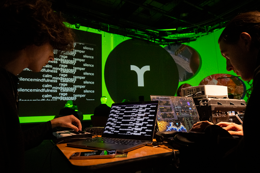 two students at a table with laptops. the laptop screens match images projected on the walls in the background