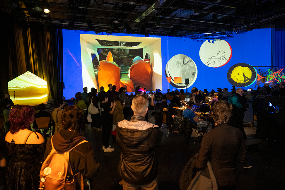 A crowd of several dozen people gathers in a dark studio space. A giant screen on the far wall shows animation projects: stop motion in a square, and several circles of hand-drawn animation