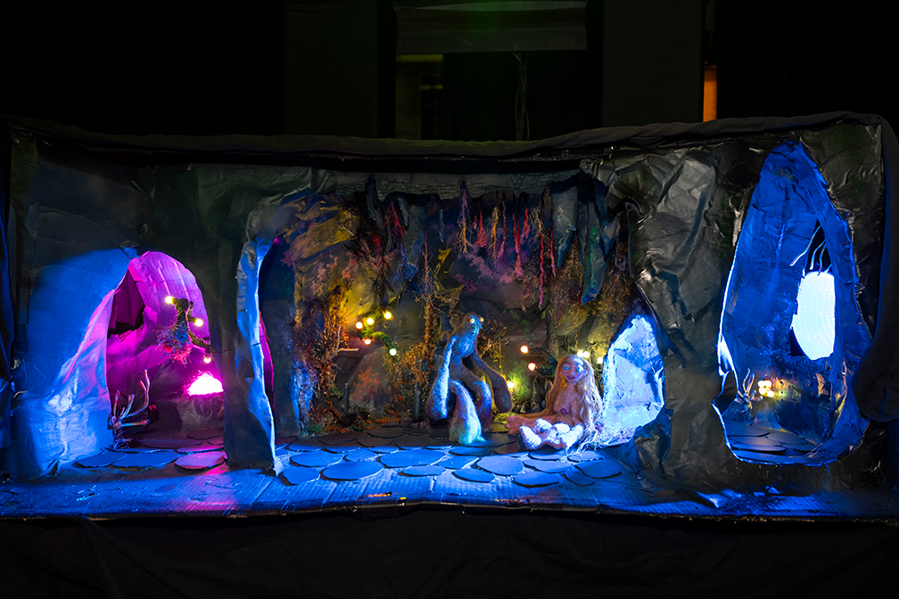 a dimly lit stop-motioin set featuring three rooms of a cave. In the middle room, a blue creature and a tan human-shaped figure with long yellow hair sit