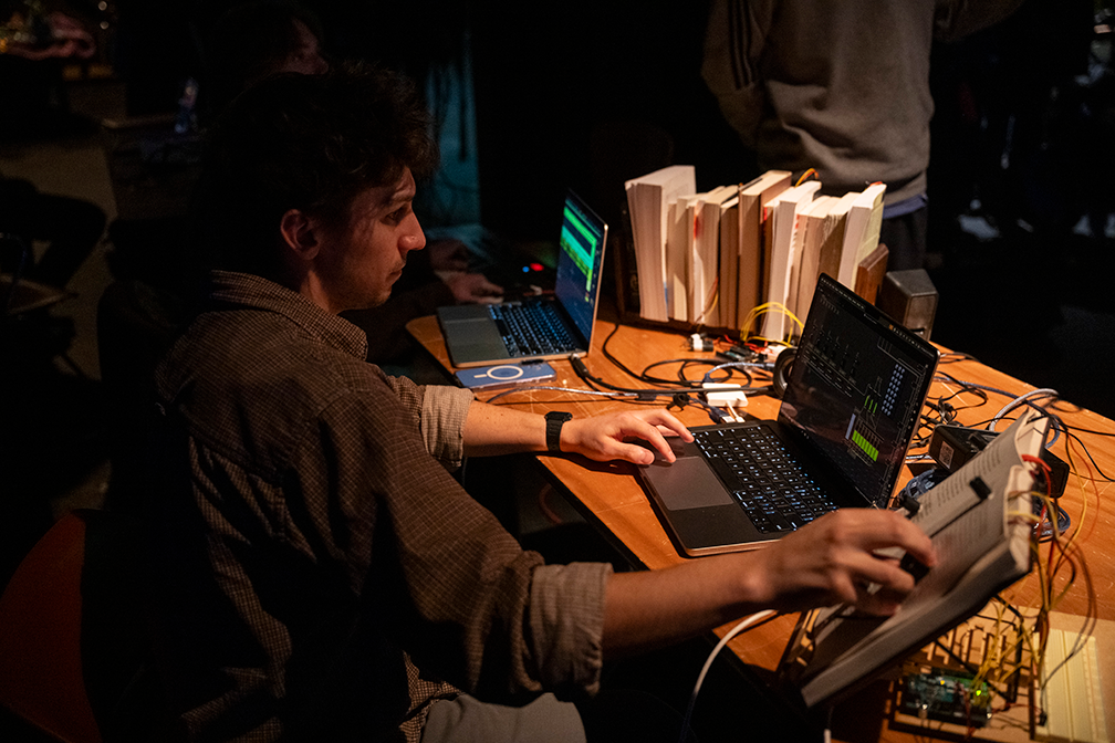 a student in a brown plaid shirt sits at a table and uses a laptop while following along in a book marked with n otes