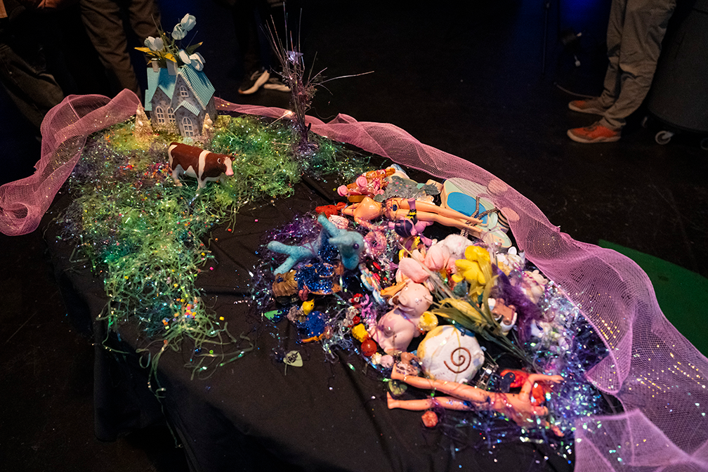 A table with a stop-motion set-up including a small plastic house, a brown and white cow, headless barbie dolls, and lots of sparkling streamers