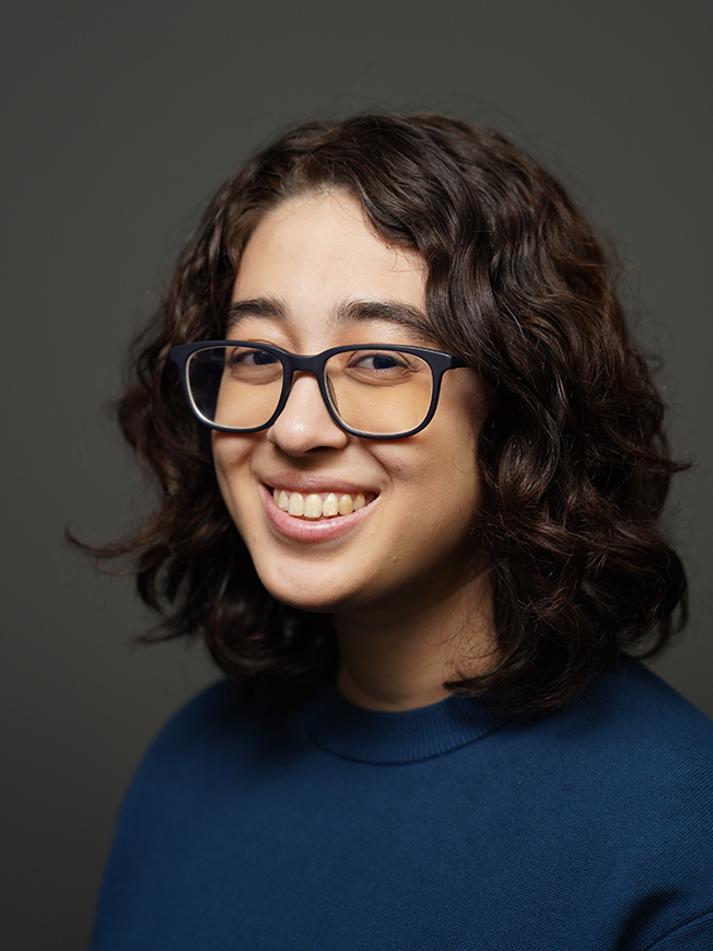 a headshot of Amanda Witt, with dark wavy hair, square glasses, and a dark blue shirt, in front of a gray background