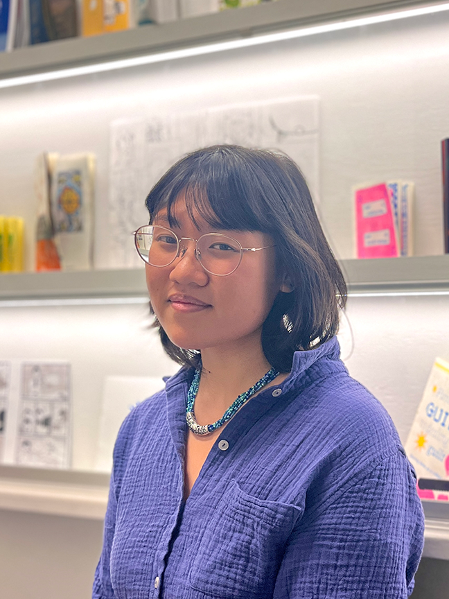 Ebba Shim, with dark shoulder-length hair, glasses, and a dark blue shirt, poses in front of a bookshelf