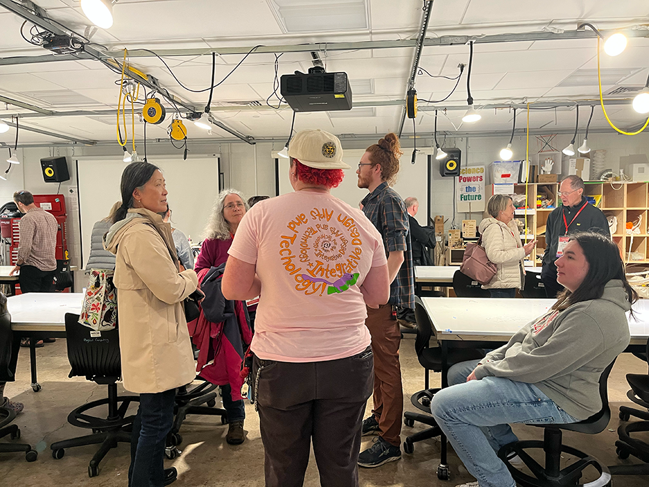 A student in a pink t shirt and white baseball cap stands with their back facing the camera, talking to six assembled people. Others mill in the background of the physical computing lab
