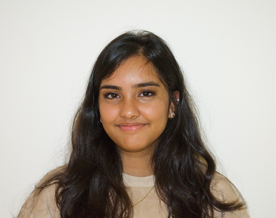 Ria Itty, with dark hair and a tan shirt, poses in front of a white background
