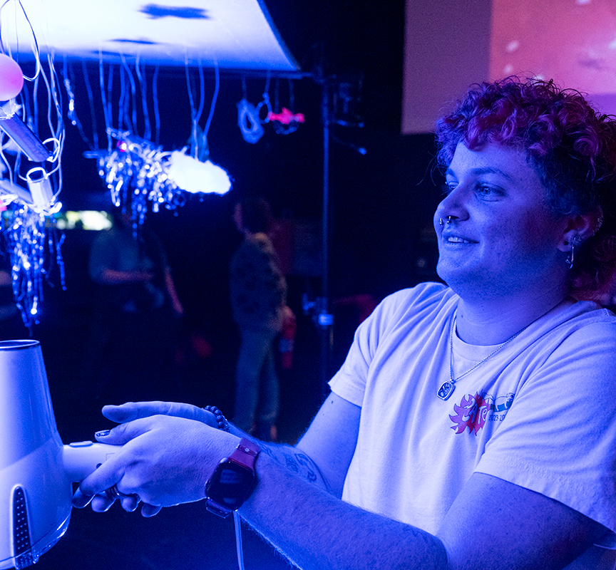 A student with short, curly hair in blue and pink lighting and a light shirt, holding a white hair dryer to interact with an exhibit