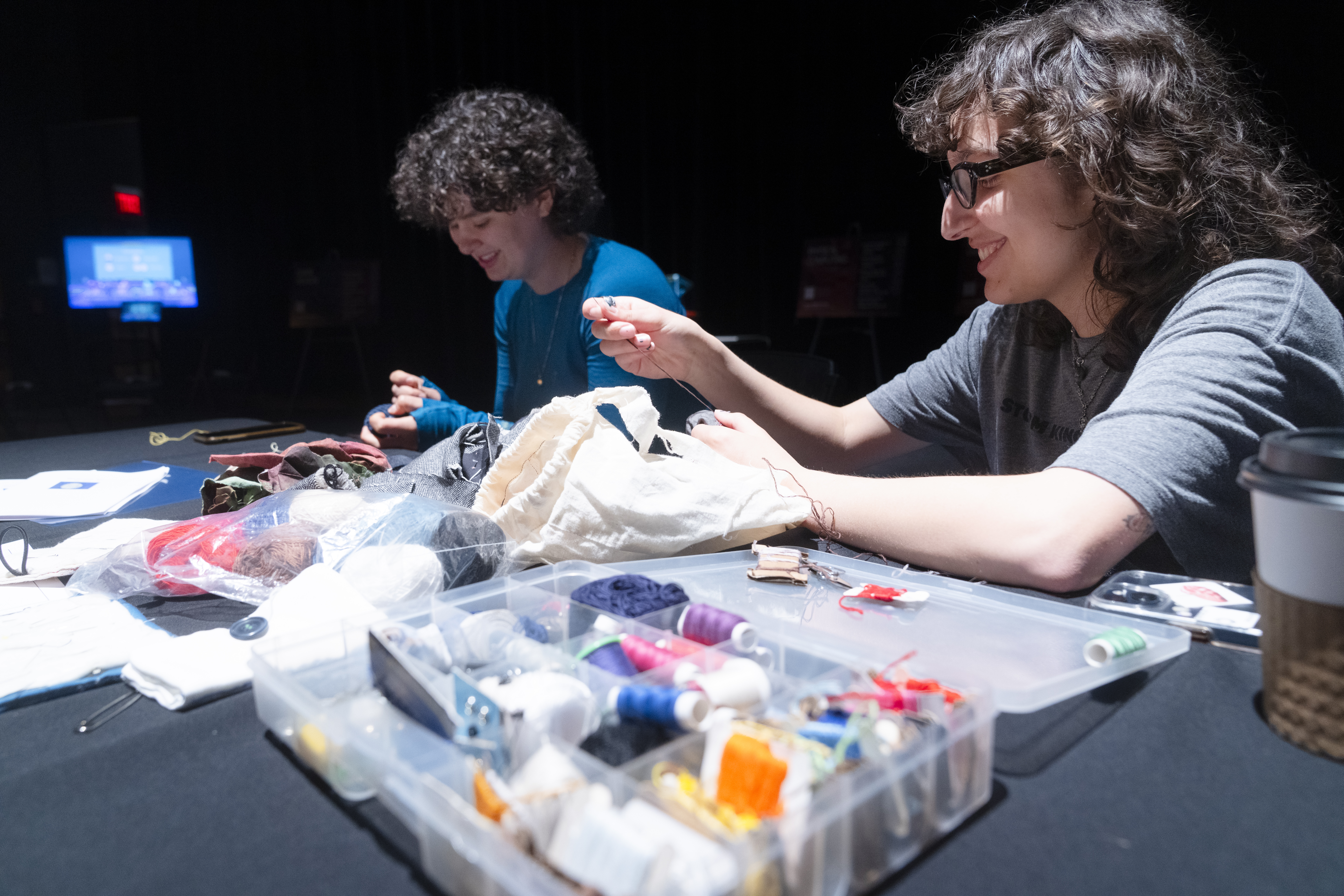 two people with short dark hair sitting at a table covered with sewing supplies. both hold thread and are mending fabrics