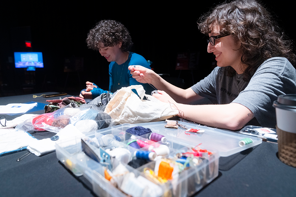 two people with short dark hair sitting at a table covered with sewing supplies. both hold thread and are mending fabrics