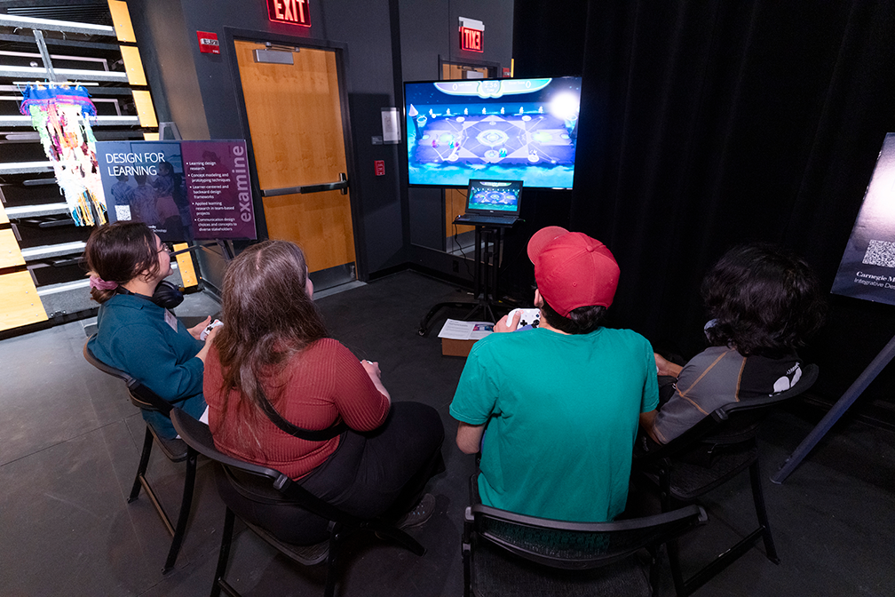 four people holding controllers gather around a large screen