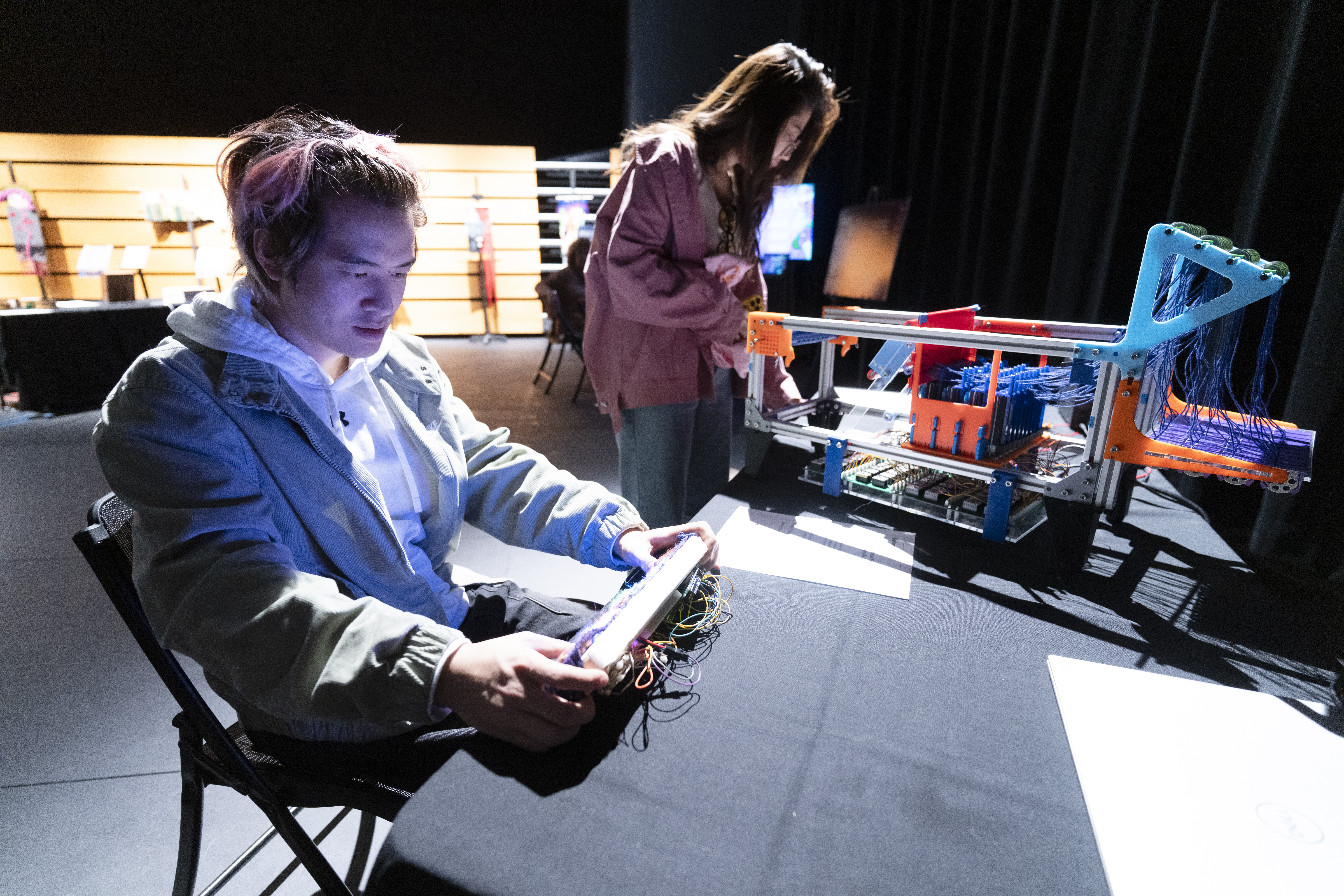 a student holds a small rectangular game console. In the background, another student views a robotic loom