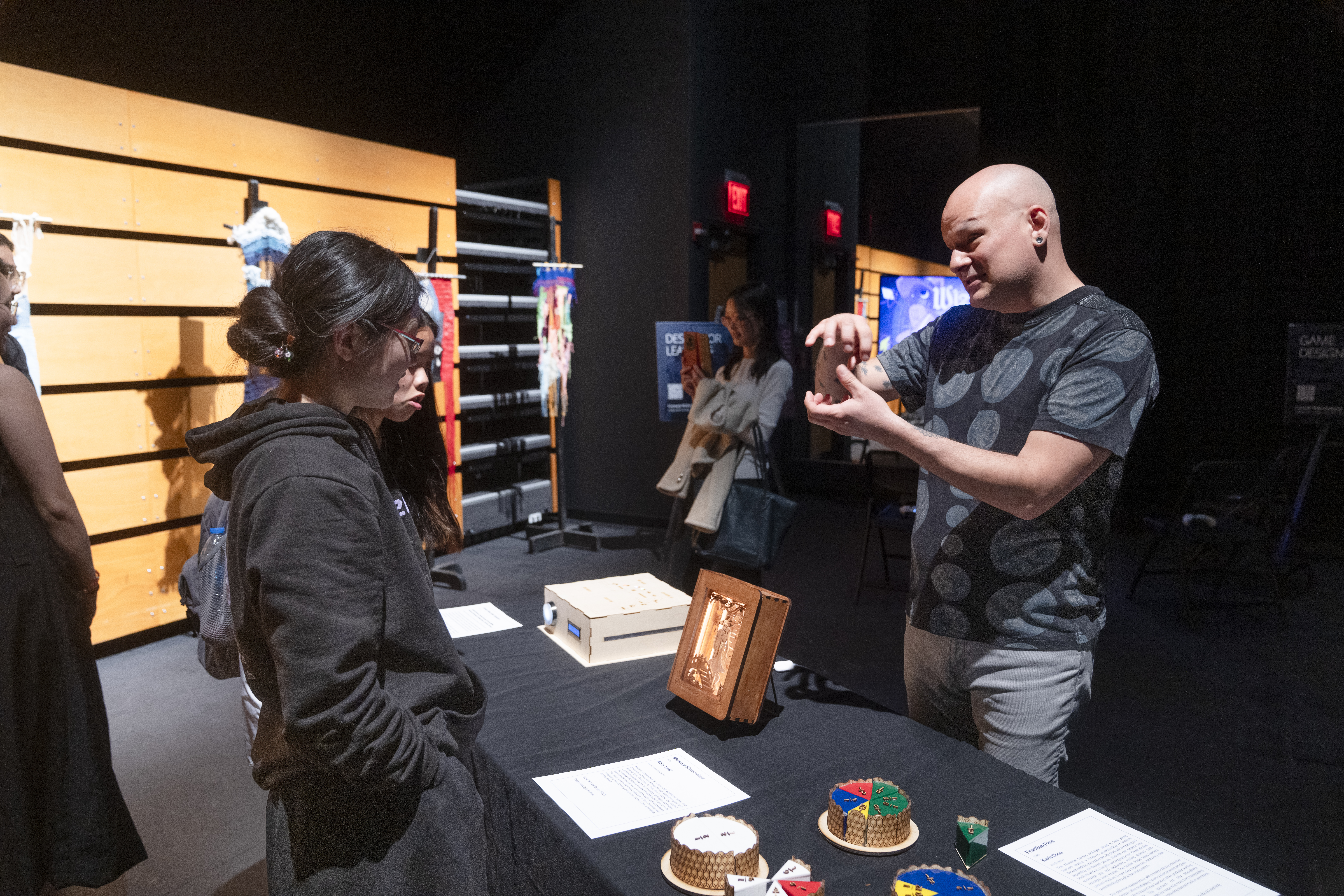 on one side of a table, students view a glowing wood box. on the other side, Ryan McKelvey uses his hands to explain the project
