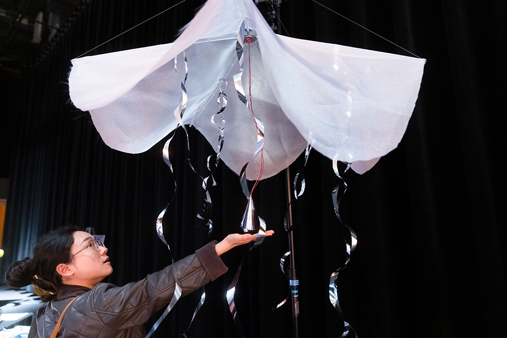 a student under a kinetic fabric sculpture titled "rain", made from 3D prints, laser cut acrylic, fabrics, strings, and sensors
