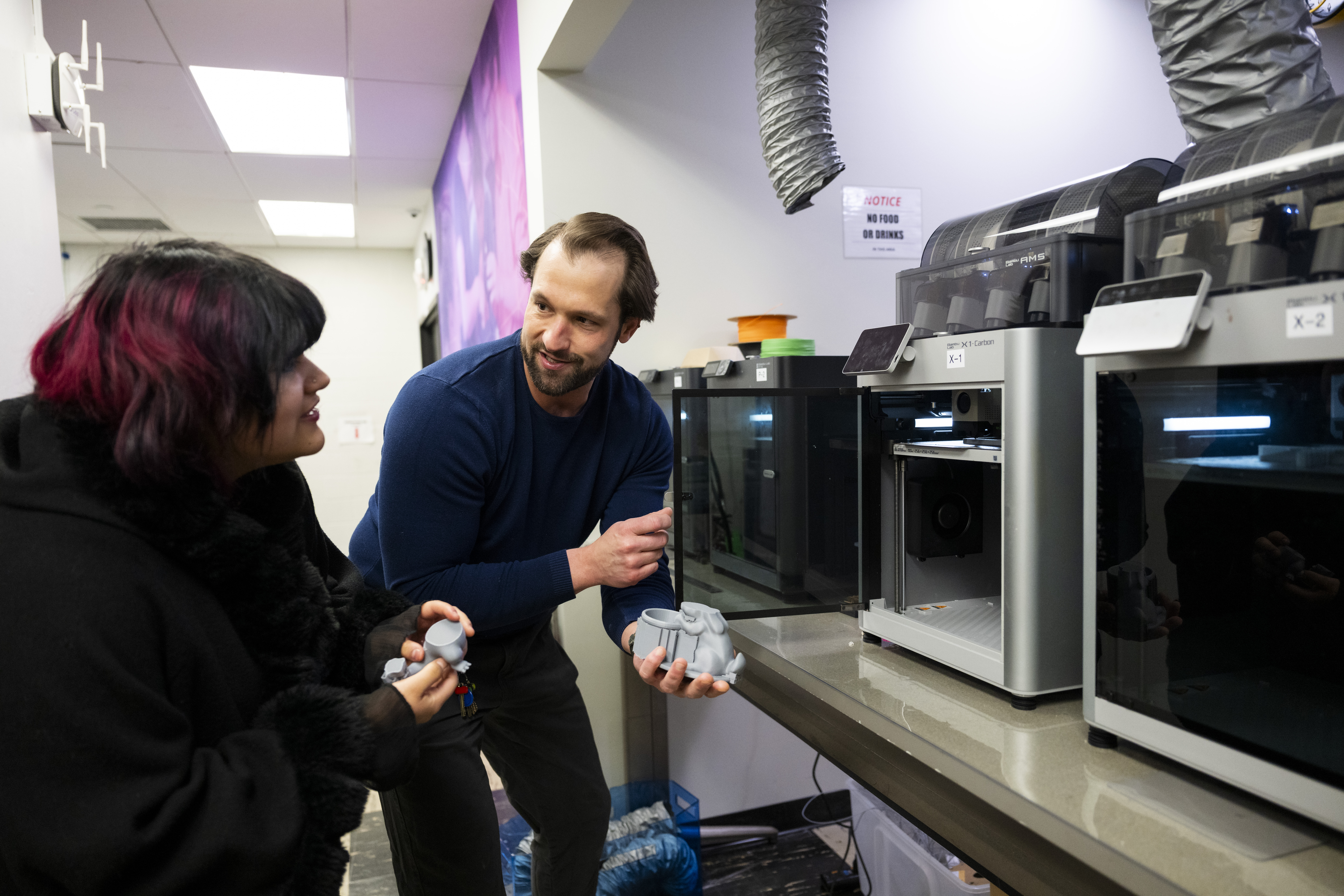 A student stands with Cody in front of a 3D printer. Both hold partial 3D printed figures in their hands.