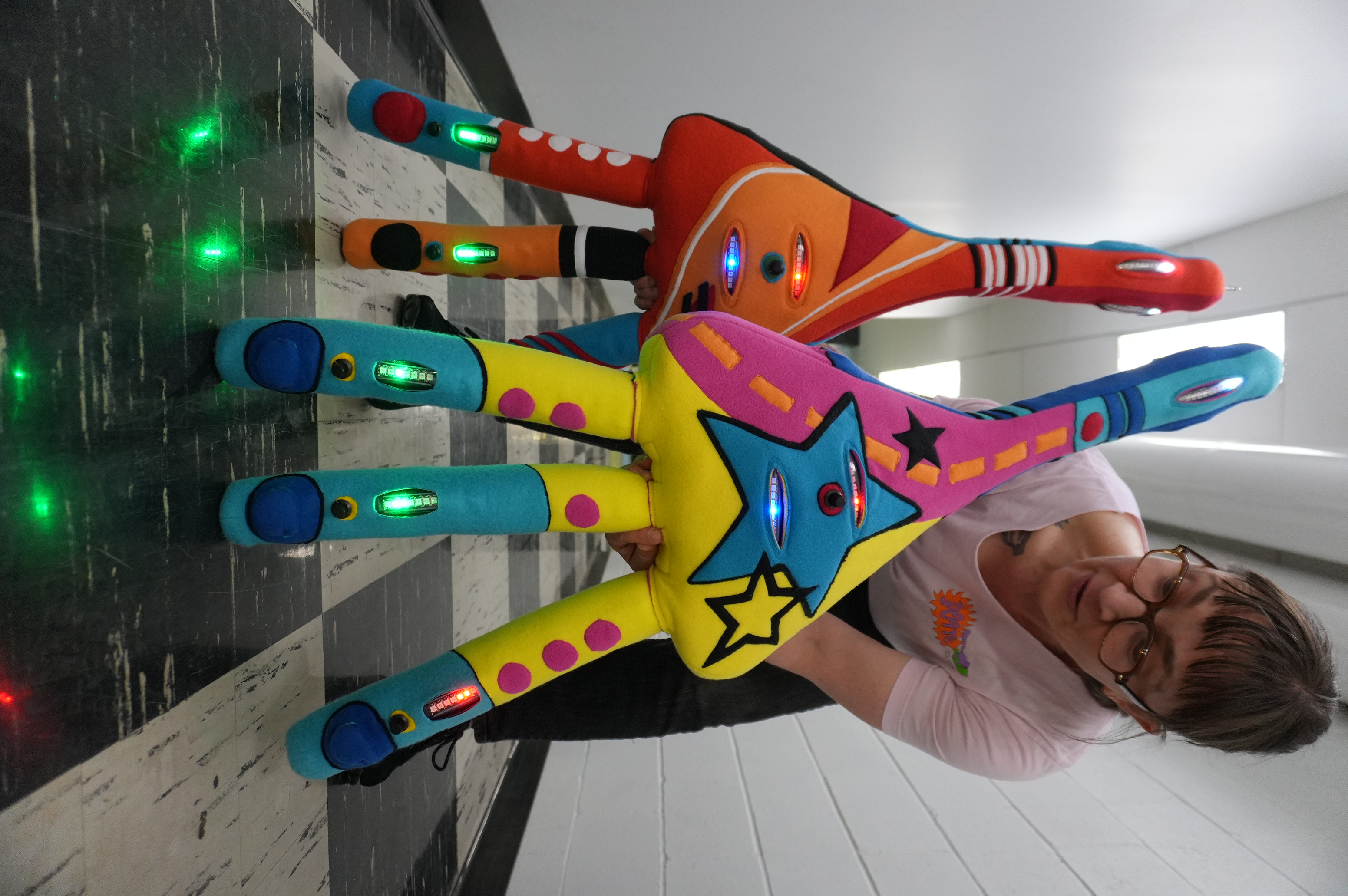 Robinson crouches in a white hallway with a black and white checkered floor, holding two Plush Neurons: one that's primarily red and orange and one that's yellow, blue, and pink