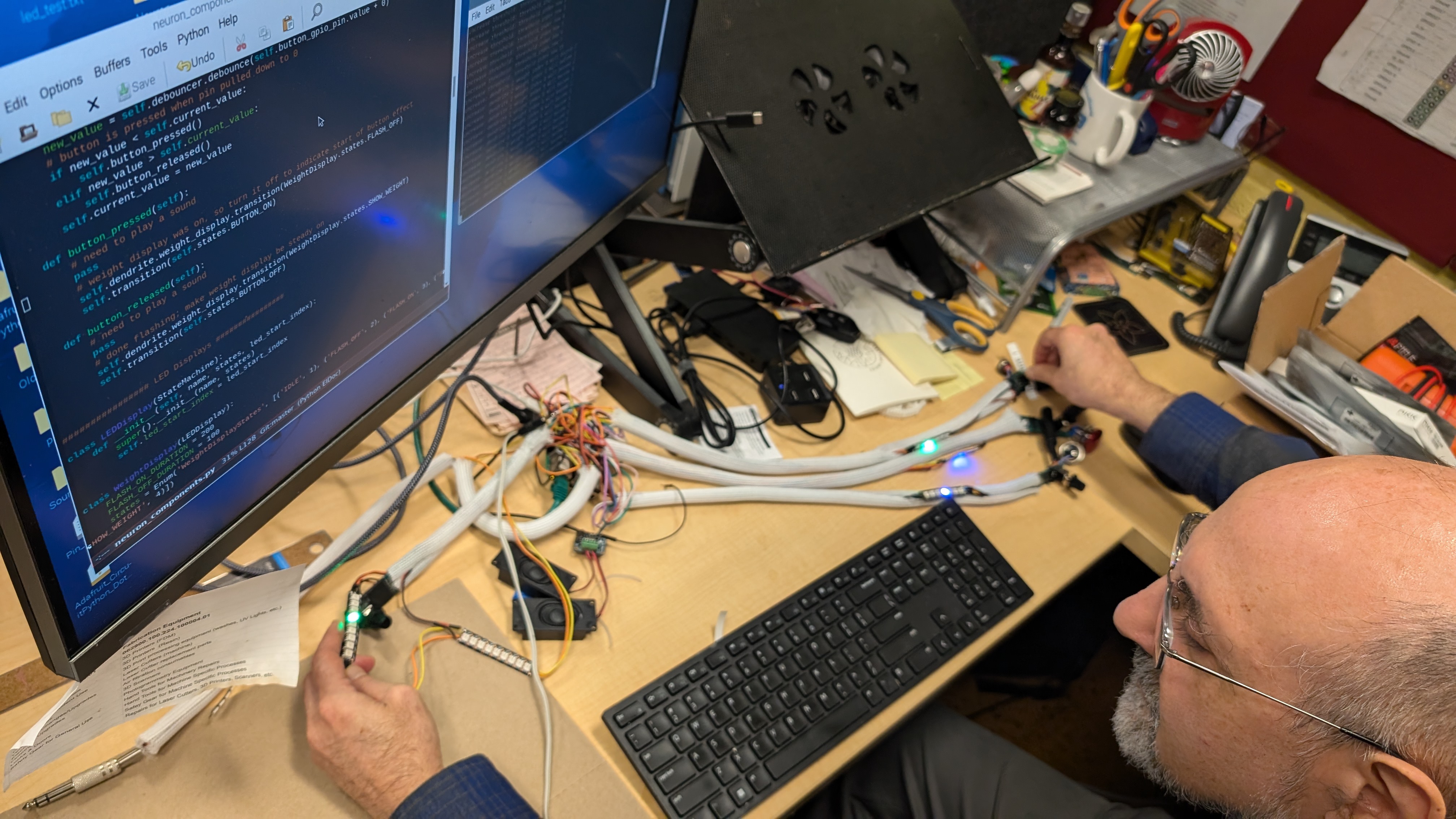 Touretzsky sits in front of a computer screen displaying code. He holds a flashing LED component in his hand that is glowing green.