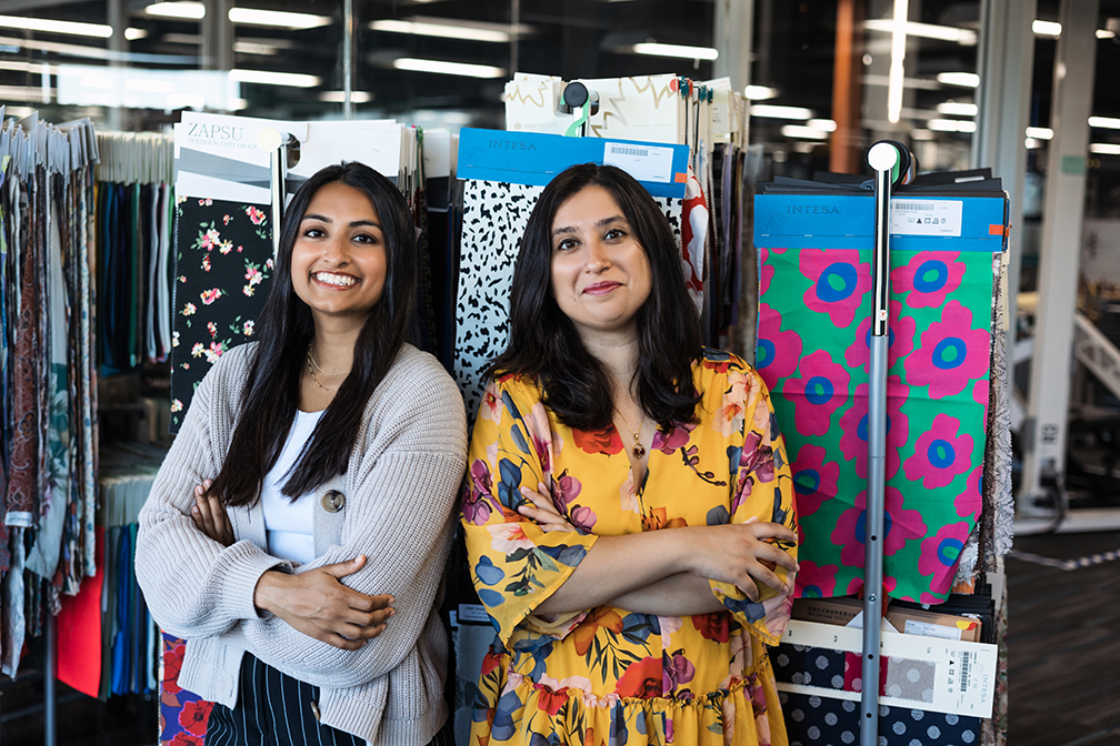 A portrait of Tushita Gupta, in a brown sweater, and Sarika Bajaj, in a yellow floral dress, standing in front of rows of hanging fabric.