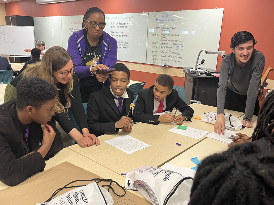 Black male middle school students in suits and ties sit around a large table, guided by their teacher in a dark purple sweater, CMU instructor Erin Gatz, and a college student in a gray long-sleeve shirt