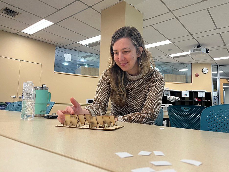 A woman sits at a table in a classroom, interacting with a laser-cut wood game with moveable pieces
