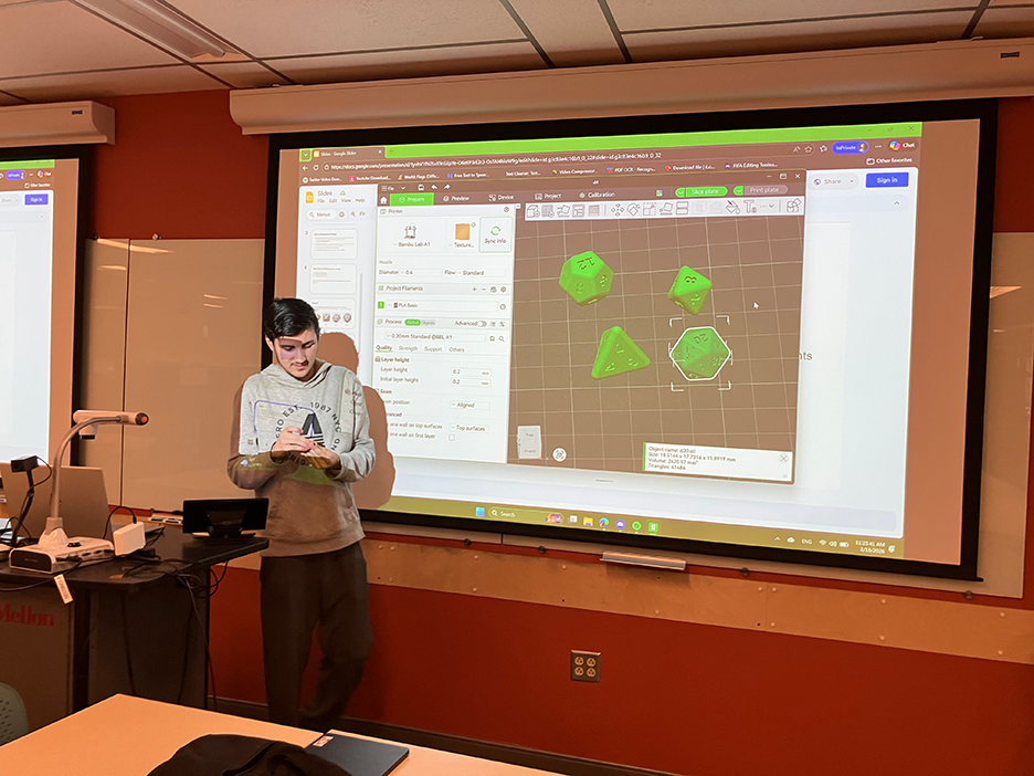 a male student stands in front of a projector screen. On the screen are designs for 3D printed dice with different numbers of sides
