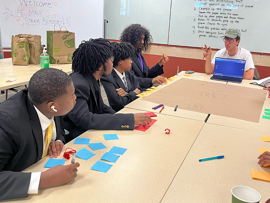 Four Black middle school boys wearing suits and ties look down the table at a college student in a baseball cap, presenting from his laptop