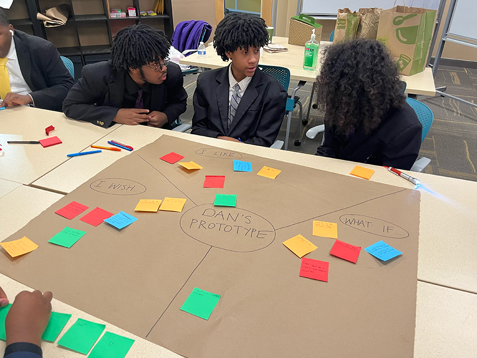 Three Black middle school boys in suits and ties discuss their feedback over a large brown sheet of paper reading "Dan's Prototype" scattered with sticky notes.