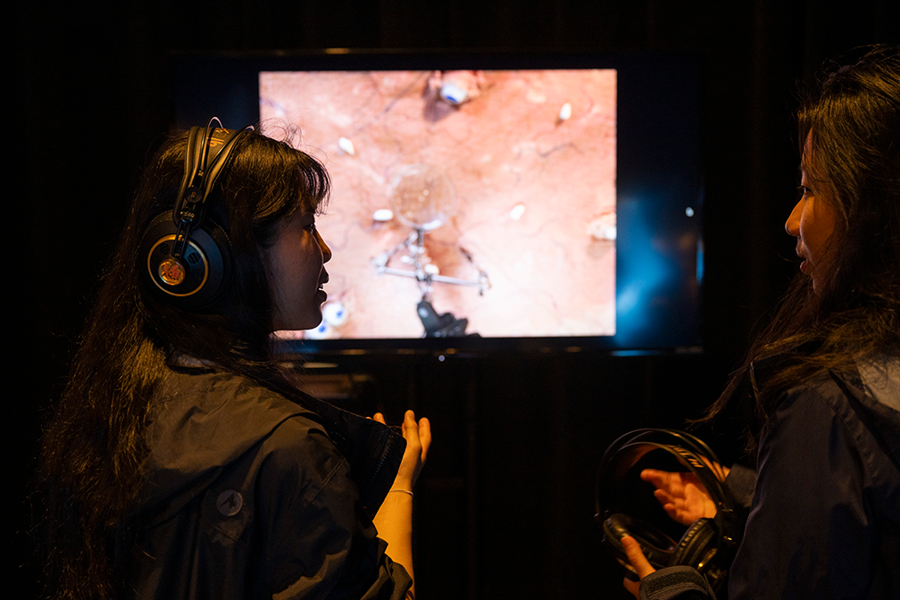 Two students, one wearing and one holding headphones, look at each other in front of a brightly lit screen