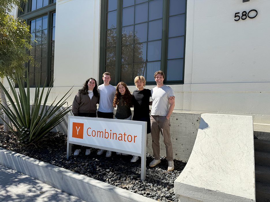 Five people in business casual clothing stand with their arms around each other behind a sign that reads "Y Combinator"