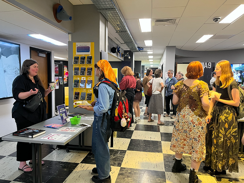 a librarian in a dark shirt and pants and sandals stands behind a table talking to a student in an orange bandana. There are books and posters across the surface of the table, and other students mingle in the background.