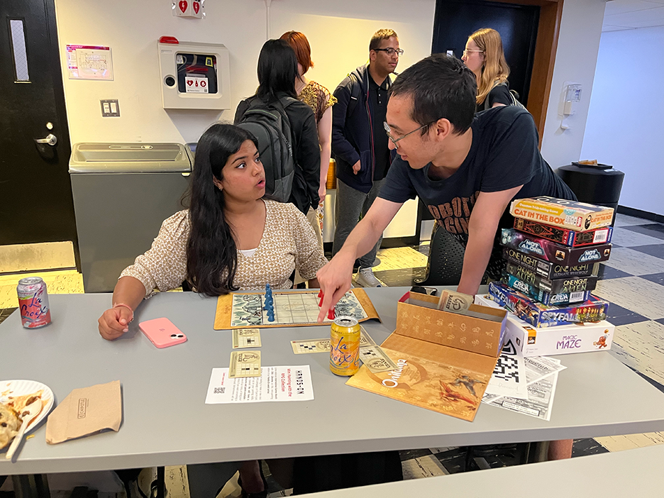 Two students, one with long dark hair and a tan shirt and the other with short hair and a black shirt, bend over a board game on a table. The game is made up of a grid of squares, and blue and red game pieces are lined up at either ends of the board.