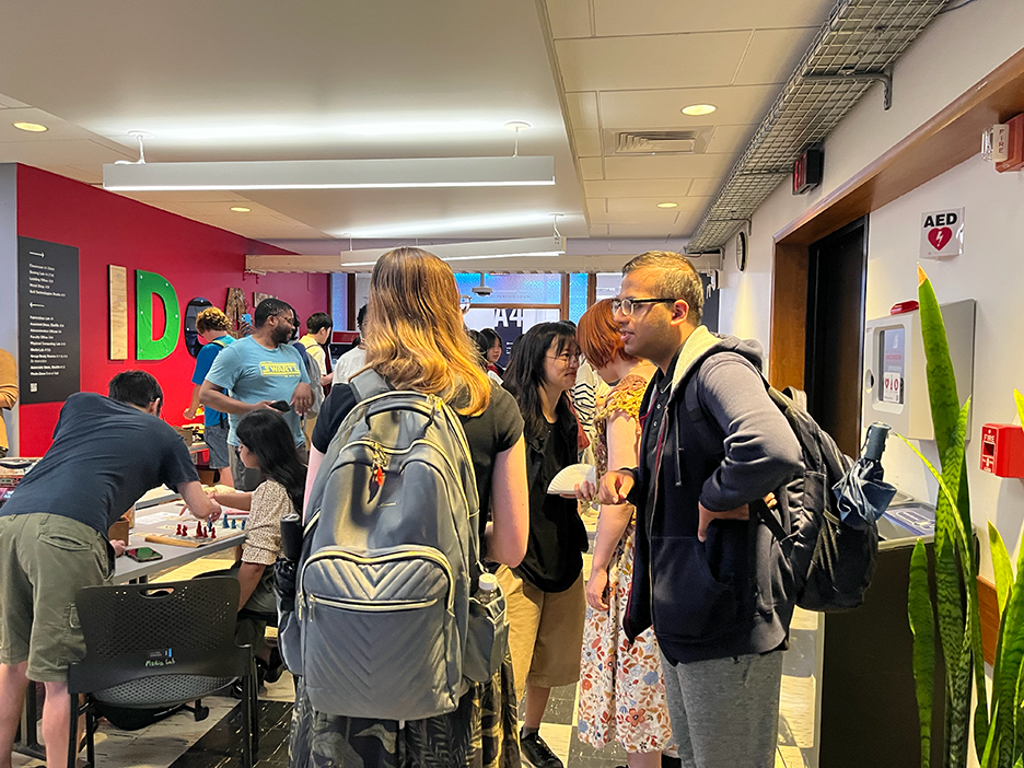 two students wearing backpacks stand in the foreground, while a crowd behind them plays games and eats burritos