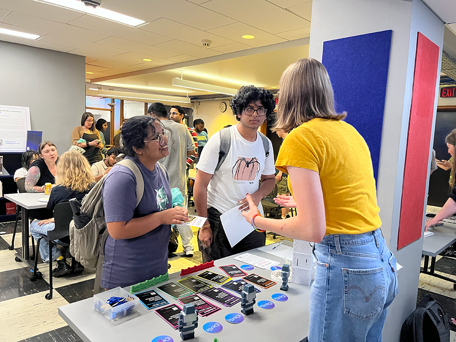 A librarian in a yellow shirt and jeans stands behind a table, and addresses two students in t-shirts and glasses.