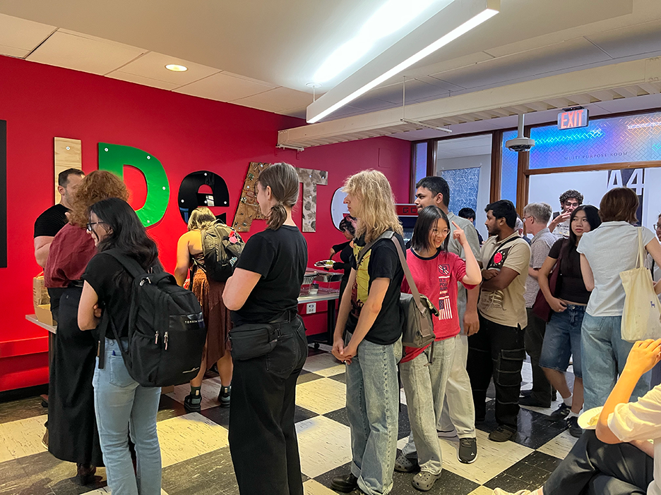 students line up along a table holding food in front of a wall painted bright red that reads "IDeATe"